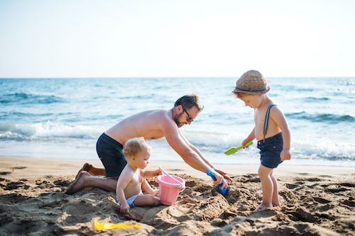A mature father with two toddler children playing with sand on beach on summer holiday.