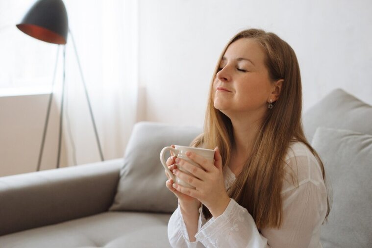 Woman sitting on couch drinking a cup of coffee closing her eyes with an optimistic and peaceful smile